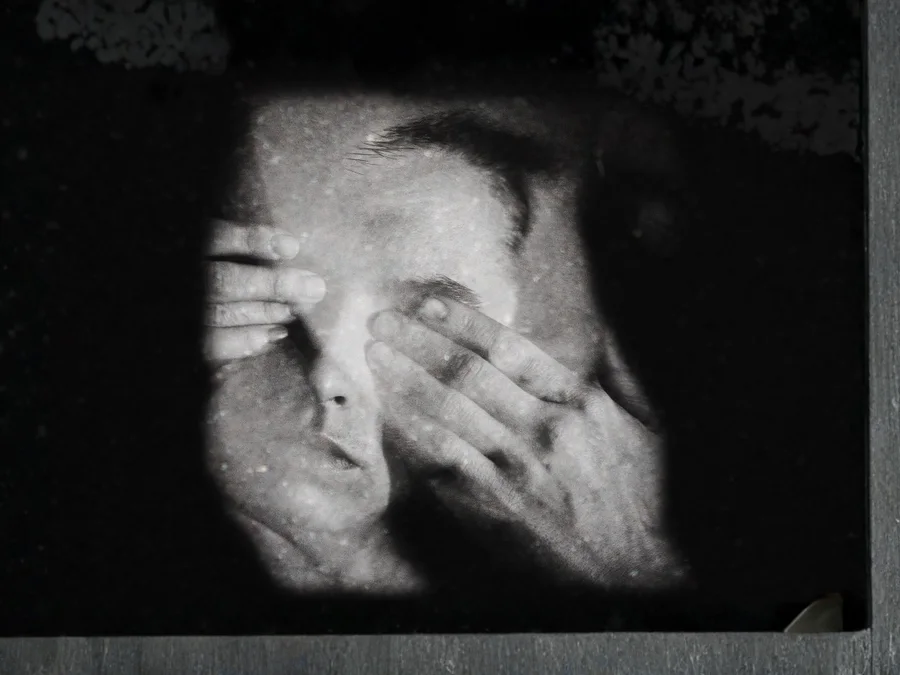 Black and white image of a person behind glass covering their eyes, suggesting workplace silence and social threat
